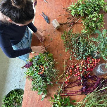 A person makes a festive garland by arranging branches, berries, and foliage on a wooden table with floral tools, twine, and scissors.