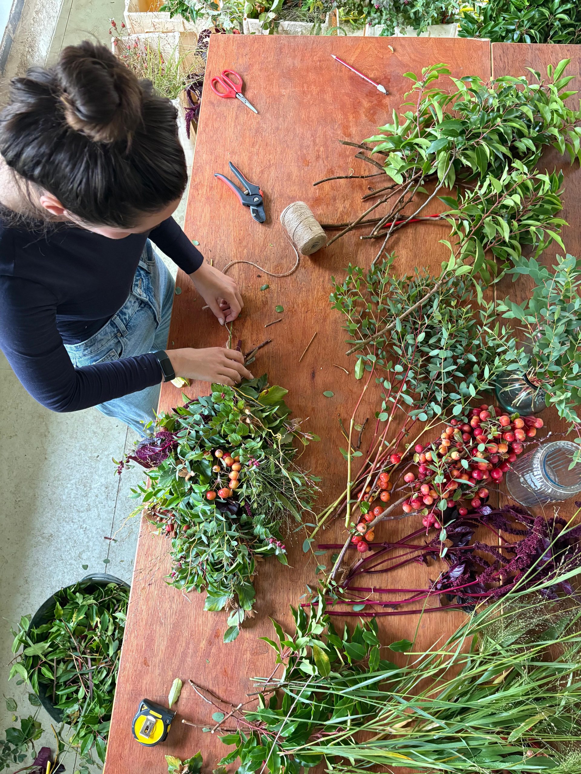 A person makes a festive garland by arranging branches, berries, and foliage on a wooden table with floral tools, twine, and scissors.