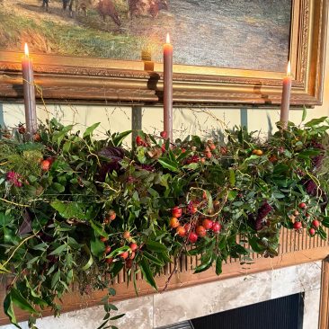 A mantel decorated with a garland from the Festive Garland Making Course, lush greenery, red berries, and three lit candles below a pastoral painting of cows.