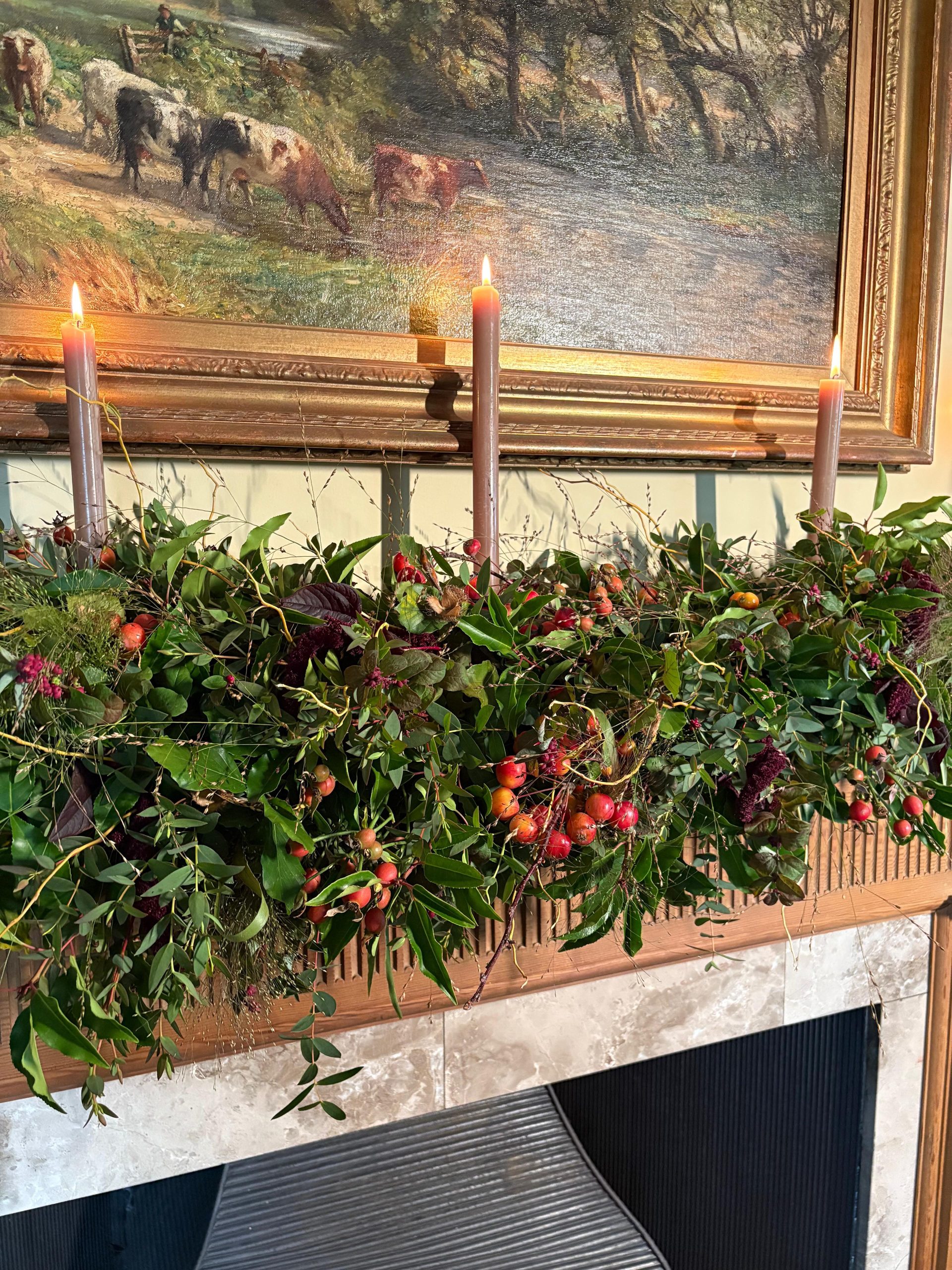 A mantel decorated with a garland from the Festive Garland Making Course, lush greenery, red berries, and three lit candles below a pastoral painting of cows.