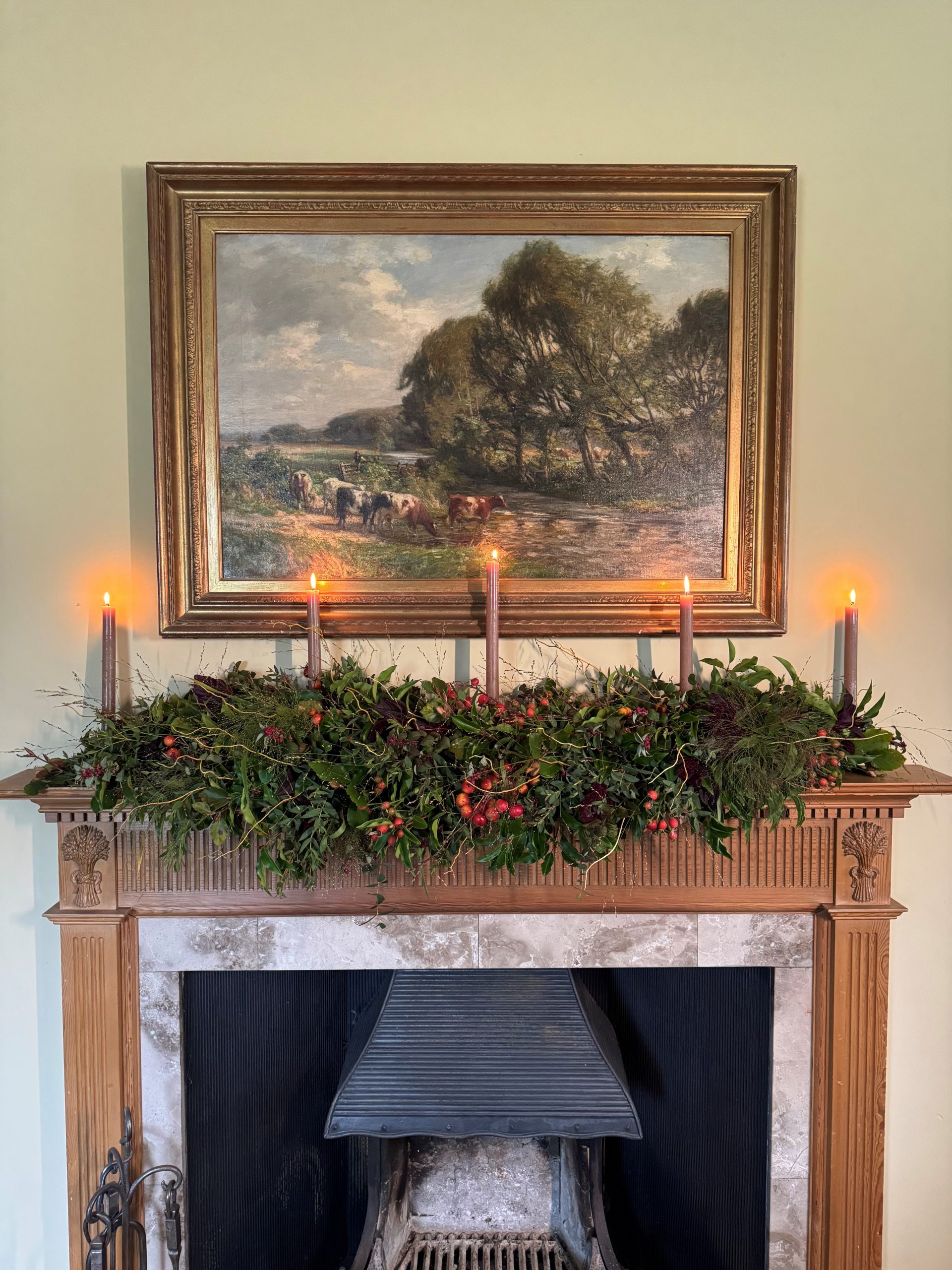 A festive mantel with lush greenery, berries and five glowing candles sits beneath a cow-filled landscape—ideal garland-making inspo.