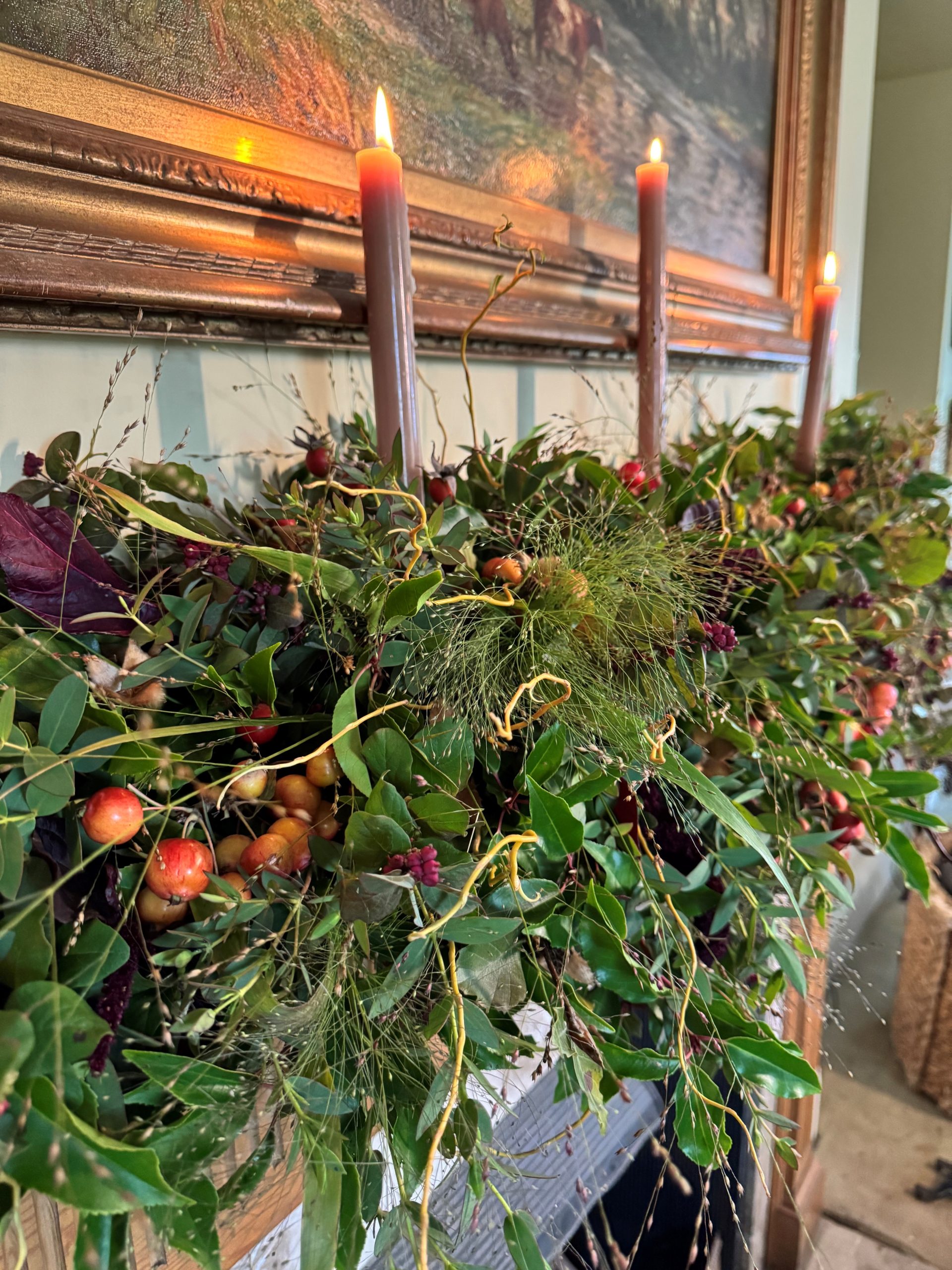 A mantel adorned with a festive garland from our course, featuring greenery, berries, and three lit candles beneath a framed painting.